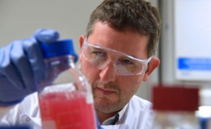 A male scientist in lab coat and goggles inspects a glass full of red liquid. 